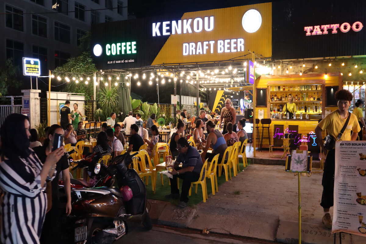 People at outdoor tables by beer sign.