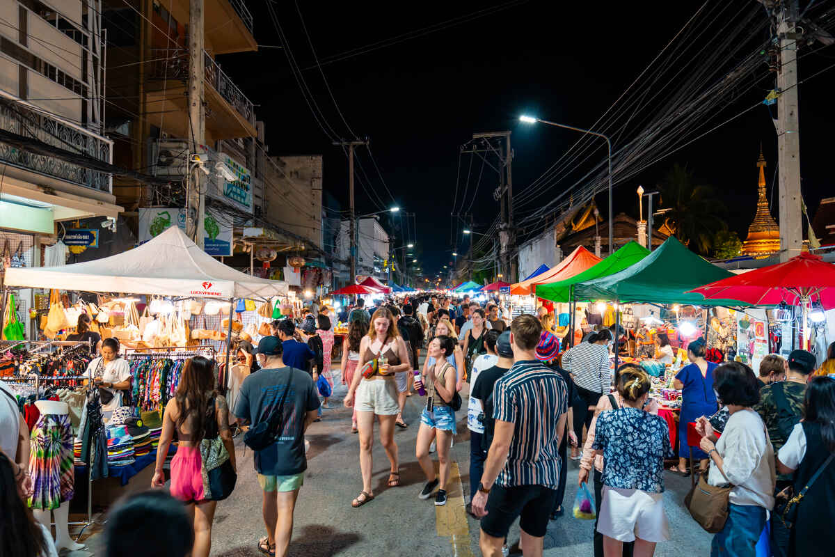 People walking through a night market with souvenirs and food and a golden Asian temple in a background