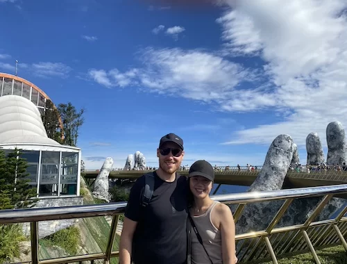 Ba Na Hills tour from Da Nang itinerary, A man and a woman smiling for the camera, standing on a bridge with similar giant stone hands in the background and a blue sky with clouds overhead.