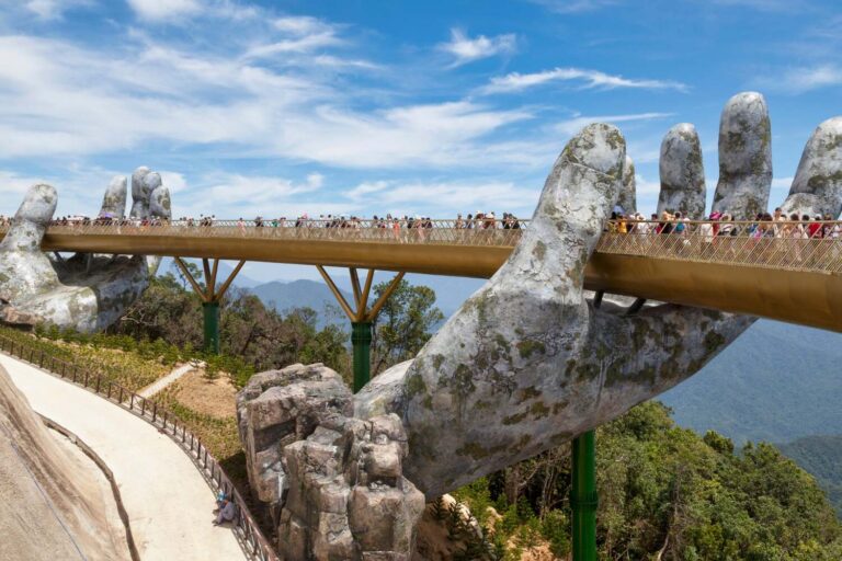 A unique bridge supported by giant stone hands extending upwards, with people walking across and greenery below.